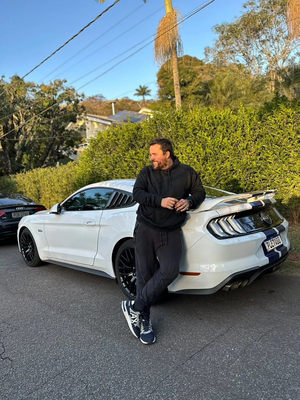 Mineiro sorrindo, apoiado em um Mustang branco estacionado na rua